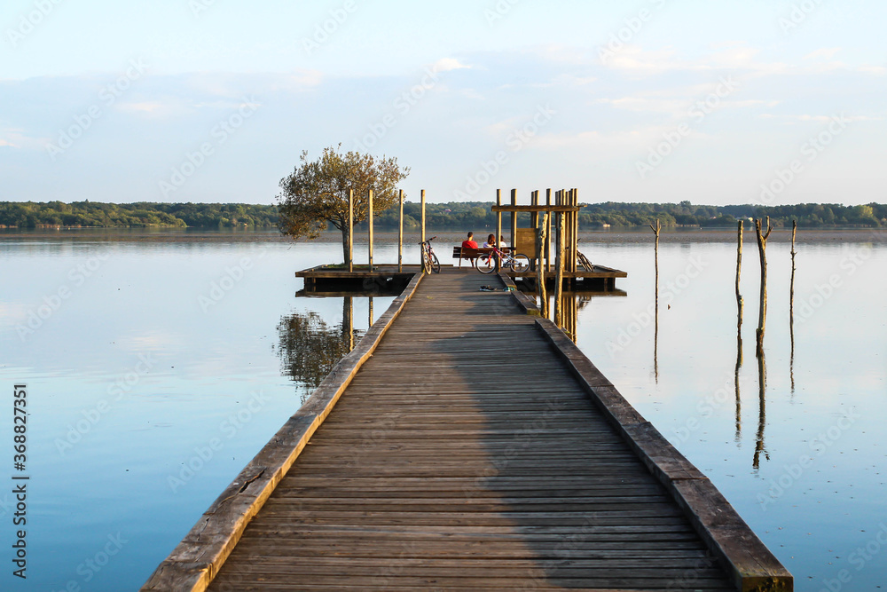 Photo de paysage : ponton avançant sur le lac avec le reflets des piquets sur l'eau. Des promeneurs sont assis au bout de la jetée et contemplent le coucher du soleil
