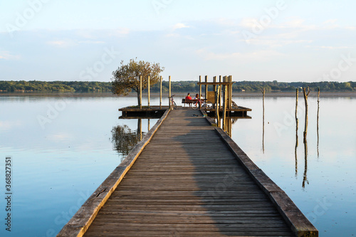 Photo de paysage : ponton avançant sur le lac avec le reflets des piquets sur l'eau. Des promeneurs sont assis au bout de la jetée et contemplent le coucher du soleil © ChabStyLe