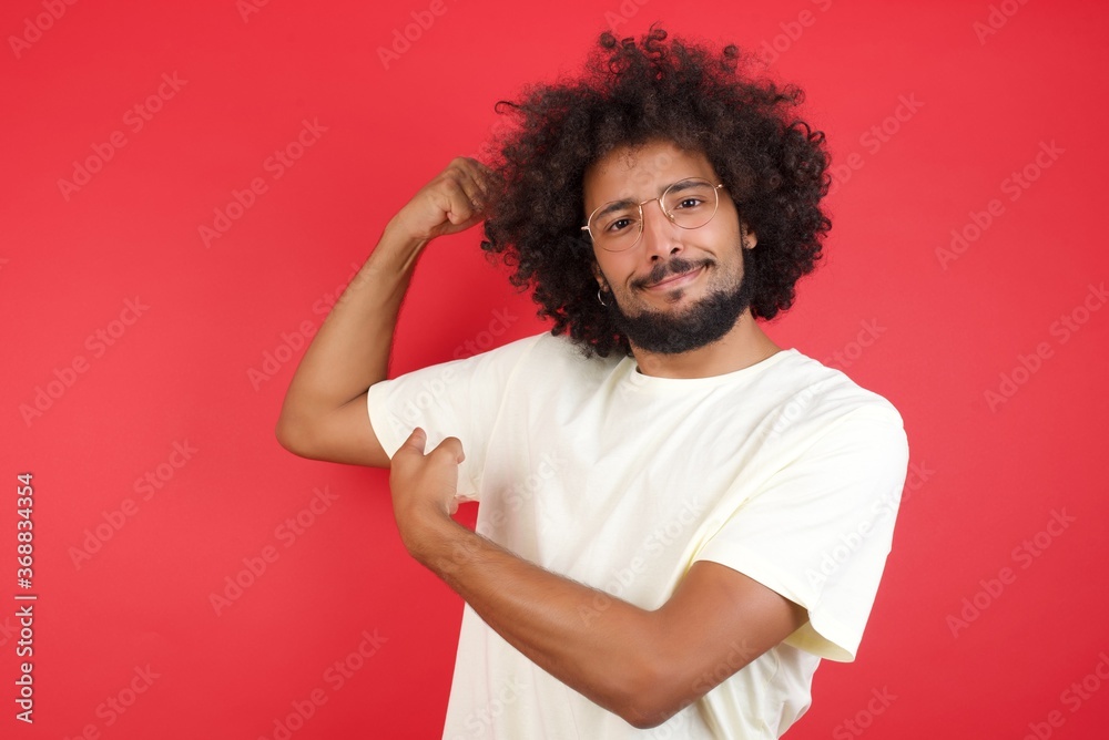 Waist up shot of blond man raises hand to show his muscles, feels confident in victory, looks strong and independent, smiles positively at camera, stands against gray background. Sport concept. Waist up shot of blond man raises hand to show his muscles, feels confident in victory, looks strong and independent, smiles positively at camera, stands against gray background. Sport concept.