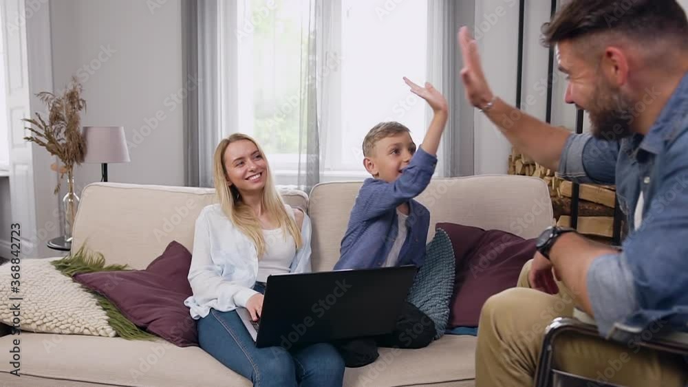 Good-natured smiling small boy giving high five to his jovial bearded father in wheelchair while browsing interesting videos on computer together with cute joyful mother at home Good-natured smiling small boy giving high five to his jovial bearded father in wheelchair while browsing interesting videos on computer together with cute joyful mother at home