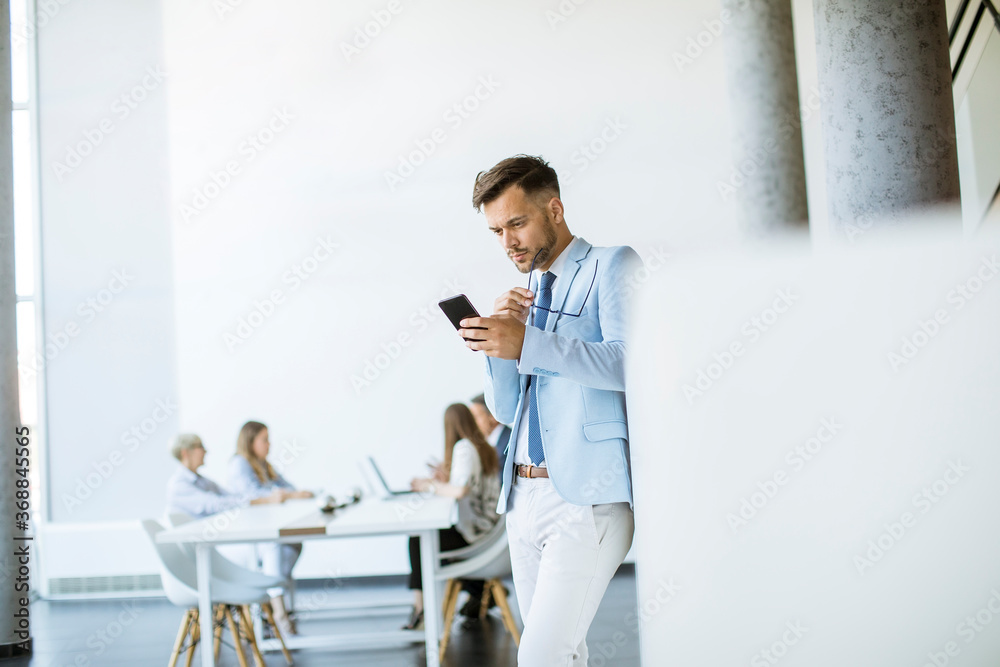 Fototapeta premium Happy young man using his mobile phone and smiling while his colleagues working in the background