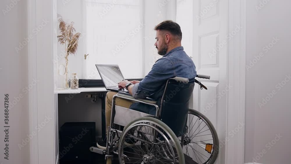 Handsome concentrated confident bearded 30-aged man in wheelchair working on laptop near window at home Handsome concentrated confident bearded 30-aged man in wheelchair working on laptop near window at home