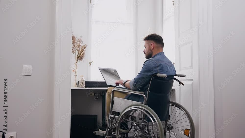 View from back attractive confident smart young bearded guy which sitting in wheelchair and remotely working on computer from his home office room View from back attractive confident smart young bearded guy which sitting in wheelchair and remotely working on computer from his home office room
