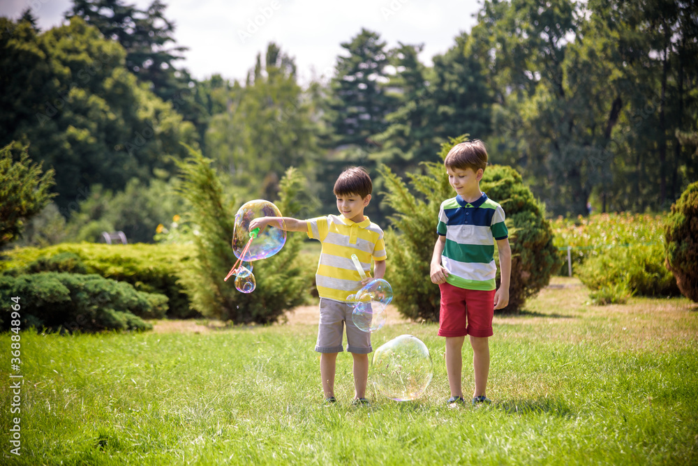 Fototapeta premium Boy blowing soap bubbles while an excited kid enjoys the bubbles. Happy teenage boy and his brother in a park enjoying making soap bubbles. Happy childhood friendship concept
