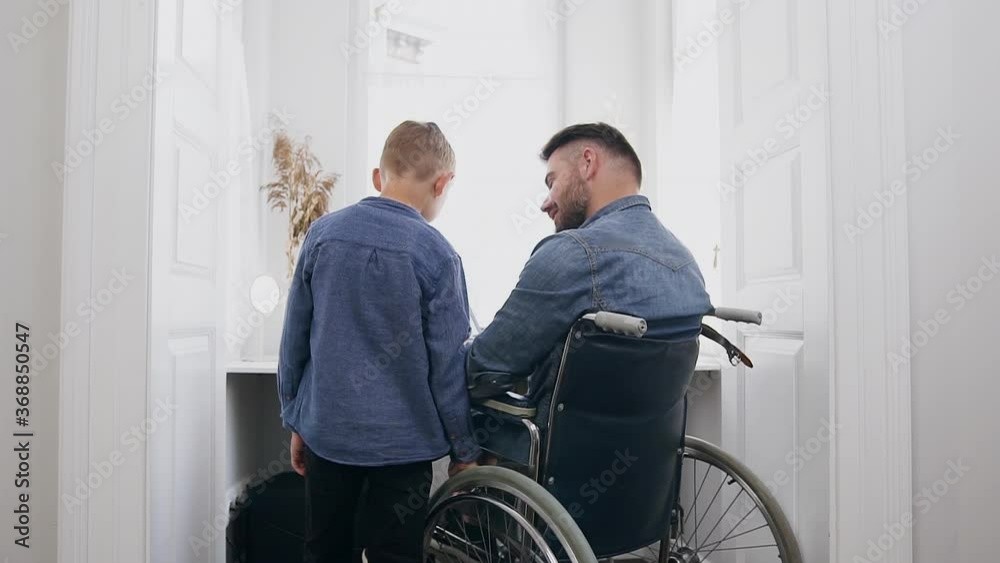 Portrait of handsome loving young bearded man in wheelchair which talking with his positive smiling 8-aged son during distantly working on computer at home Portrait of handsome loving young bearded man in wheelchair which talking with his positive smiling 8-aged son during distantly working on computer at home