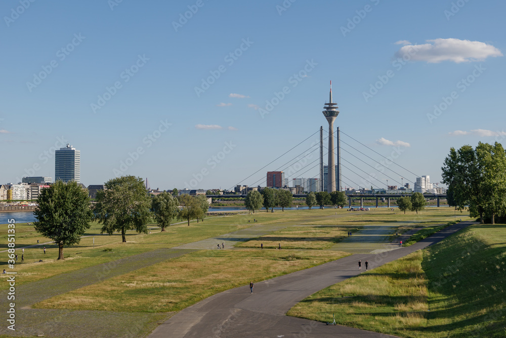 Aerial outdoor panoramic sunny scenic landscape view of natural promenade and green field on riverside of Rhine River, and background of skyline cityscape of Düsseldorf city, Germany in summer season.