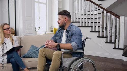 Attractive sedentary young bearded man sitting in wheelchair and talking with attantive female medical assistant during she visiting him at home photo