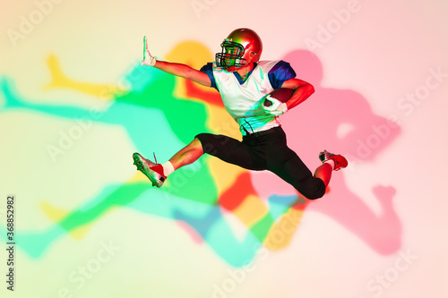 American football player isolated on gradient studio background in neon light with shadows. Professional sportsman during game playing in action and motion. Concept of sport, movement, achievements.