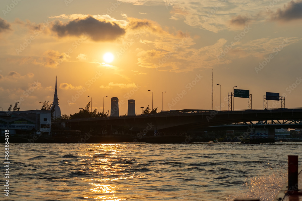 Sunset silhouette of temple, buildings and bridge from boat on Chao Phraya river in Bangkok, Thailand  