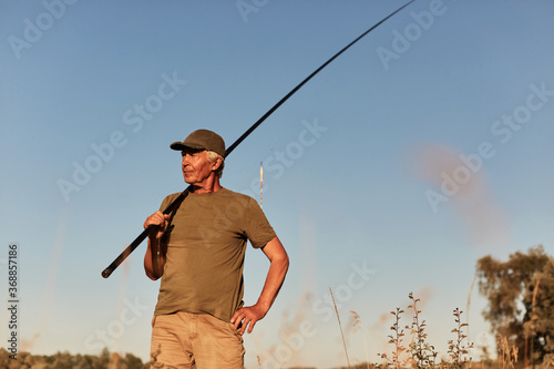 Fisherman posing with fishing rods in hands with blue sky on background, being ready to catch fish, looking pensive side, wearing green t shirt and short.
