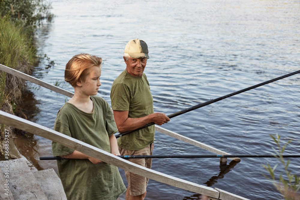 Father and son fishing on wooden stairs with rods in hands, dad looking at his boy with love, family spending time together in open air, guys wearing green t shirts. Father and son fishing on wooden stairs with rods in hands, dad looking at his boy with love, family spending time together in open air, guys wearing green t shirts.