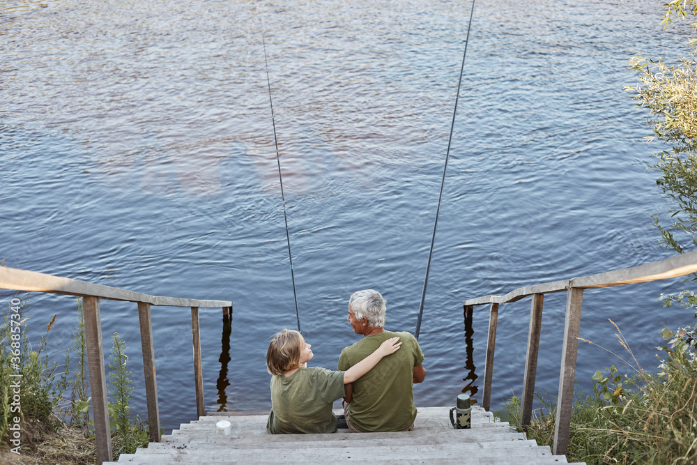 Fototapeta premium Happy family spending time together in open air near river or lake, son hugging his father with love while sitting on wooden stairs leading to water.