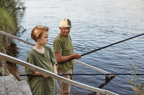 Father and son fishing on wooden stairs with rods in hands, dad looking at his boy with love, family spending time together in open air, guys wearing green t shirts.