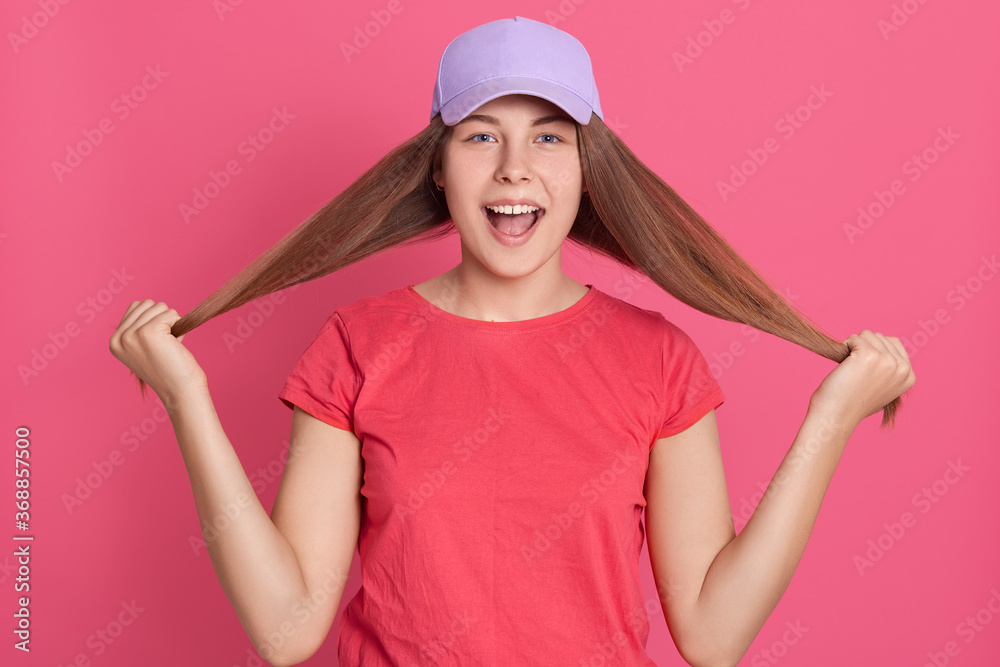 Positive young lovely female pulling her long straight hair with raised hand and looking cheerfully directly at camera, isolated over pink background. Positive young lovely female pulling her long straight hair with raised hand and looking cheerfully directly at camera, isolated over pink background.