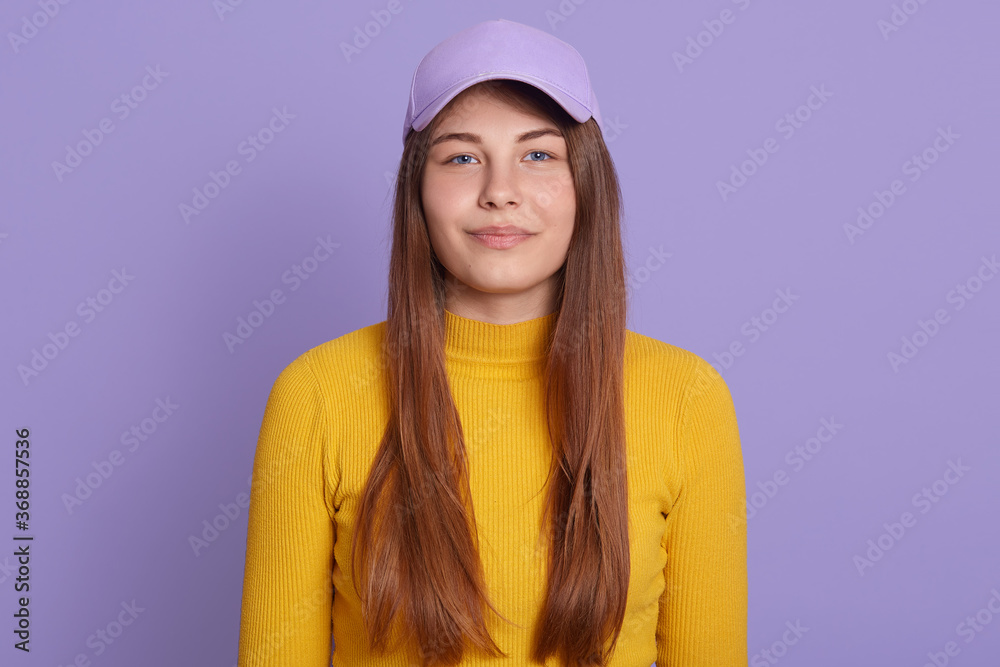 Close up portrait of tender charming girl, posing smiles, standing in baseball hat isolated over lilac background, wearing yellow casual shirt, being in good mood.