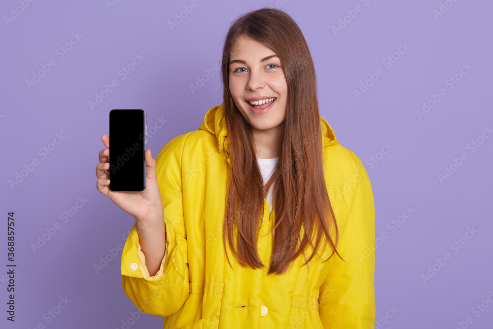 Girl showing blank smart phone display and looking directly at camera with happy smile, posing isolated over lilac background, wearing yellow shirt. Girl showing blank smart phone display and looking directly at camera with happy smile, posing isolated over lilac background, wearing yellow shirt.