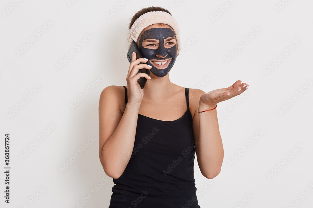 Portrait of beautiful woman during spa treatment at home while talking via modern smart phone, spreading palm aside, looking smiling aside, posing isolated over white background.