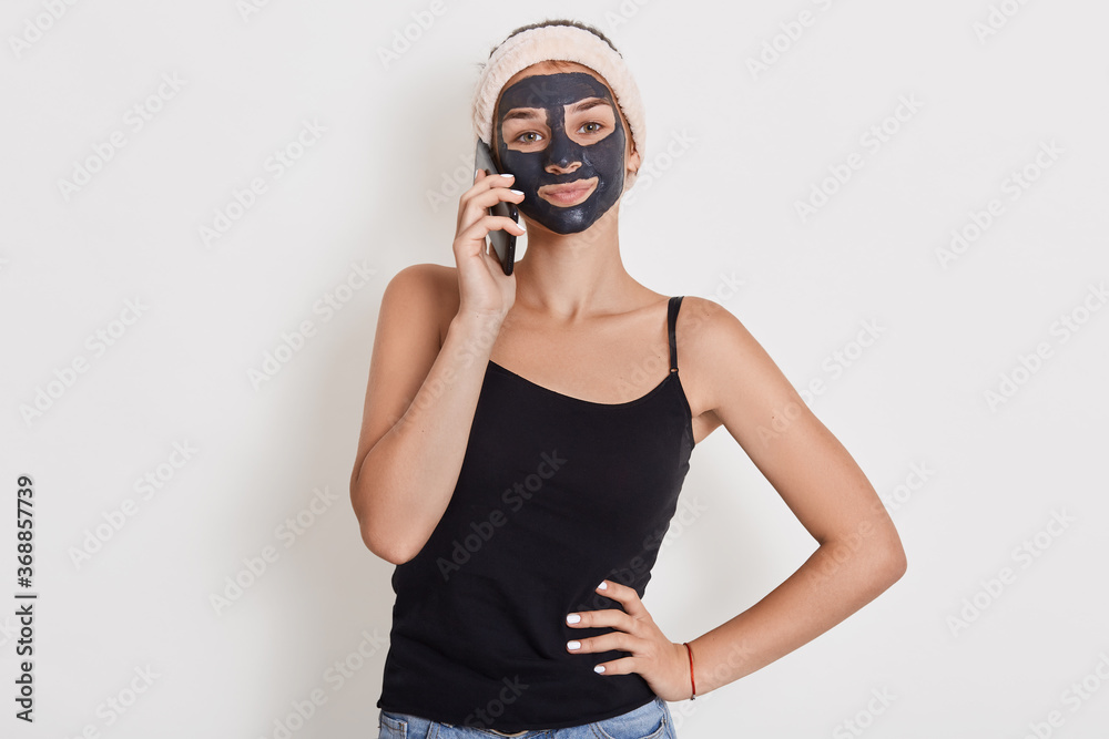 Young woman with hair band on head and black facial clay mask, talking via phone. Spa beauty treatments, skin care at home, happy woman against white wall.