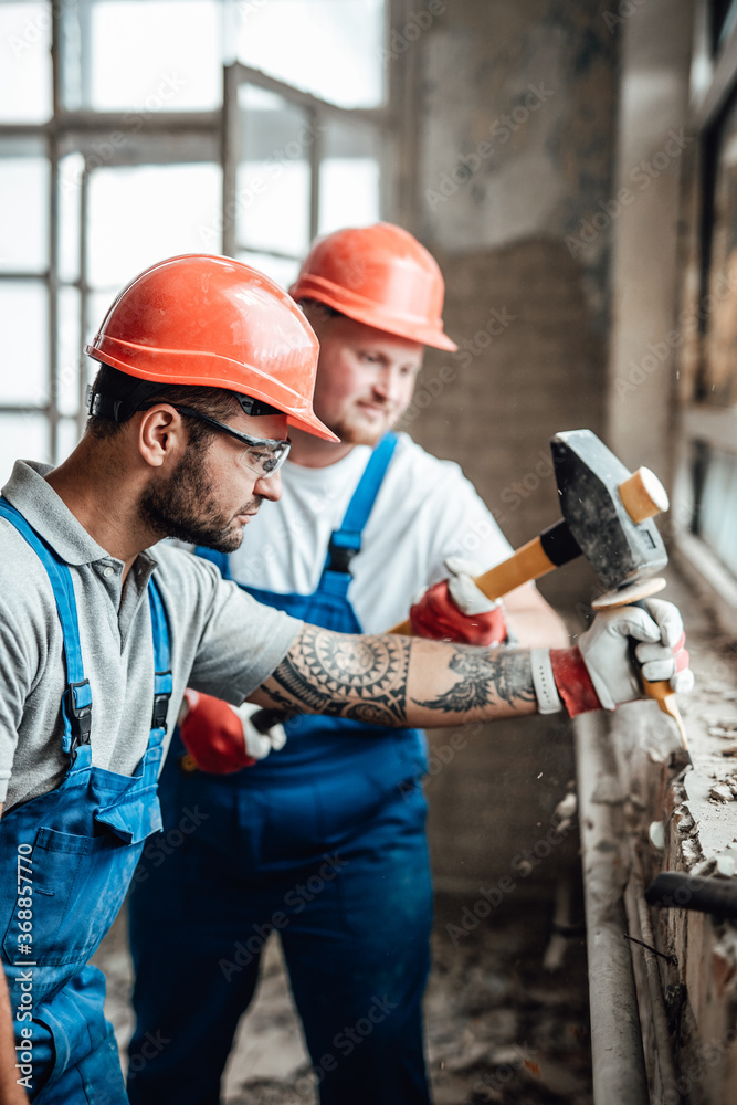 A couple of workers at a large construction site removing plaster from an old brick wall near a window