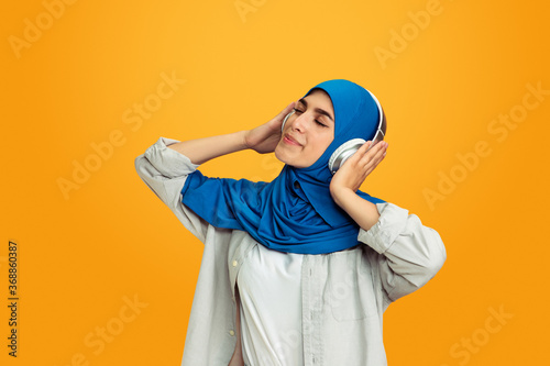 Joyful listening to music with headphones. Young muslim woman on yellow background. Stylish, trendy and beautiful female model. Human emotions, facial expression, sales, ad, shopping concept © master1305