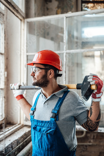 Worker, taking a short break, stands near the window at a construction site. He is wearing a red safety helmet and he holds a sledgehammer in his hands.