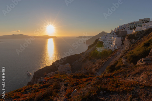 Santorini - view of the rocky coast overgrown with heather and low trees. In the background white houses illuminated by the setting sun and beautiful sea with islands.