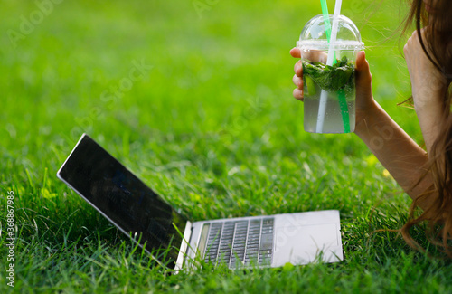 Image of girls hand with a lemonade works with a laptop in the park on a green lawn.