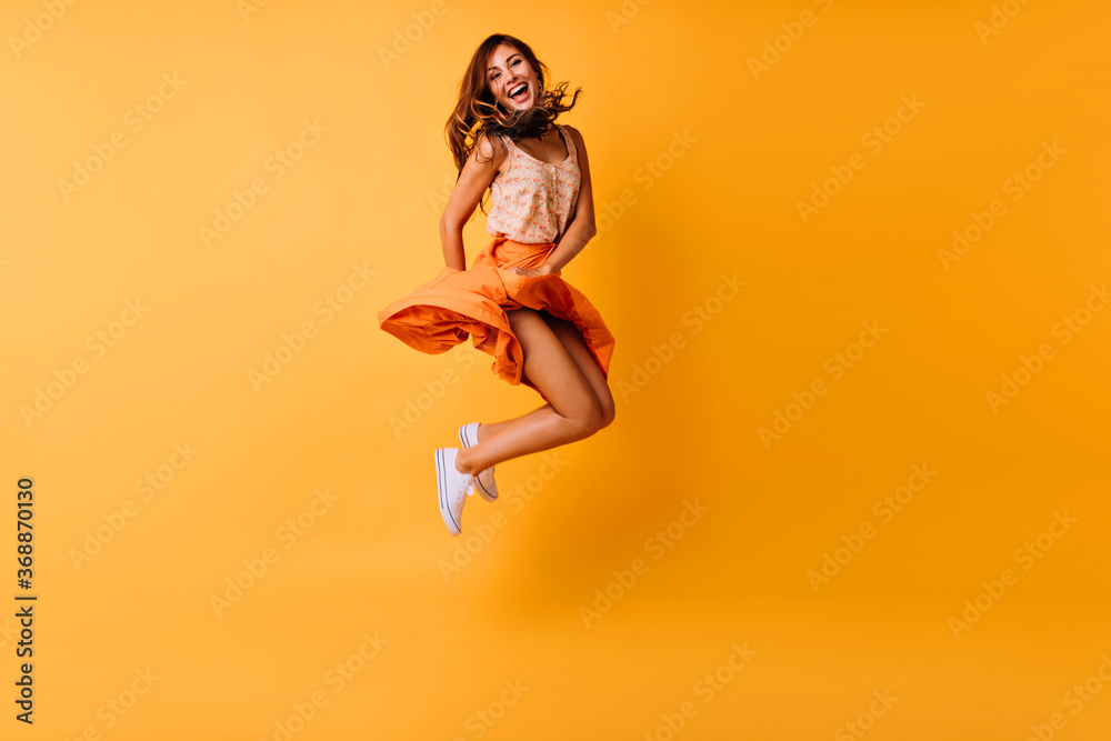 Full-length photo of tanned ginger girl jumping on yellow background and smiling. Adorable young woman in trendy summer clothes fooling around in studio.