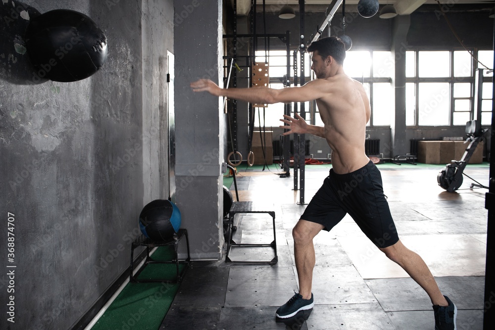 Young strong sweaty focused fit muscular man doing throwing medicine ball up on the wall for crossfit training hard core workout in the gym