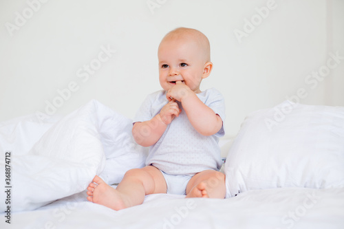 Cute baby boy 6 months smiling in a white bodysuit sitting on a bed on white bedding