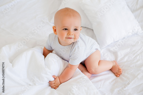 Cute baby boy 6 months smiling in a white bodysuit sitting on a bed on white bedding