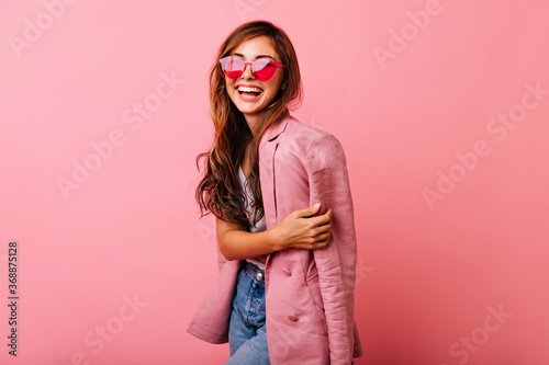 Wonderful young woman with long hair having fun on rosy background. Magnificent girl in trendy sunglasses relaxing during photoshoot. photo