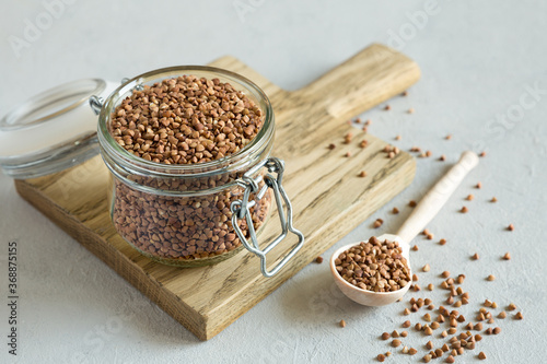 Buckwheat uncooked grains in glass jar on the wooden cutting board on gray background close up. Wooden spoon with grits. Healthy, dietary food. Organic, carbohydrate product. Food storage.