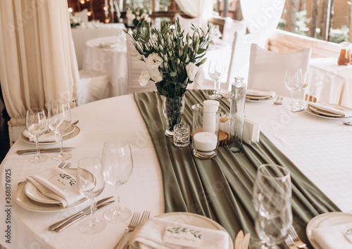 glass vase with a bouquet of white flowers in the middle of the served festive table. olive colore drapery on a white set table © Tetiana
