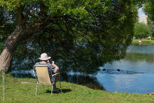 Elderly woman in a white hat sits on a folding chair near a pond in a city park and solves a crossword puzzle on a sunny summer day