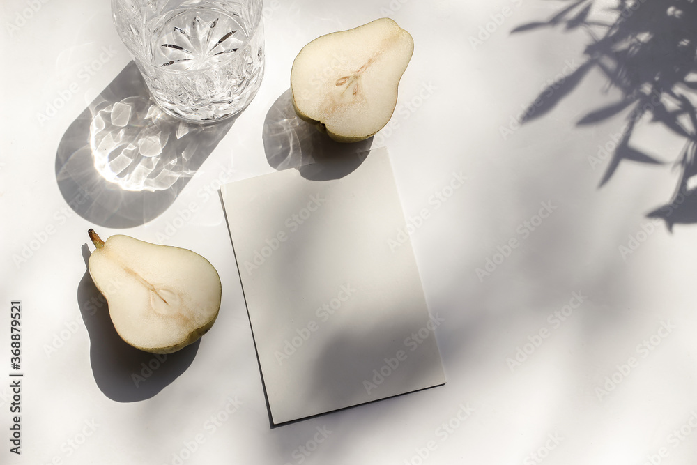 Summer stationery still life scene. Glass of water and cut pear fruit in sunlight. White table. Blank paper card, invitation mockup scene, olive branches shadows. Flat lay, top view, no people. - obrazy, fototapety, plakaty 