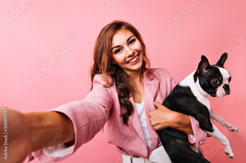 Stunning girl with cheerful smile making selfie with bulldog. Studio shot of blissful european lady fooling around with puppy.