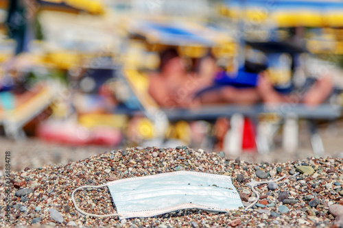 Discarded used facial mask lies on a sandy pebble beach, in the background beachgoers are relaxing by sun loungers and beach parasols. Becici, Budva Municipality, Montenegro