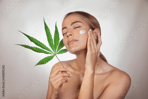 Woman applying CBD facial cream made from cannabis extract for a natural skin treatment. Portrait of young woman with cannabis leaf. Cosmetology and treatment concept. Isolated on gray background
