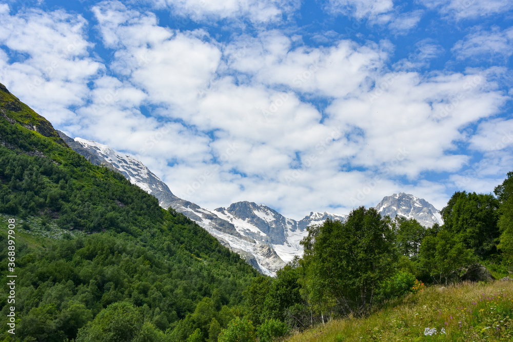 A beautiful view of the snow-capped mountain peaks of the Caucasus Mountains. White clouds against the blue blue sky.