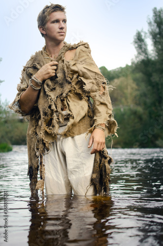 medieval peasant in a ragged raincoat of old sacking wades through the river photo