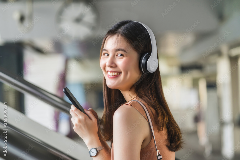 Young Asian woman passenger wearing headphone and using smart mobile phone to listening music and walking up the stairs in subway station when traveling in big city, commuter and transportation concep