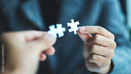 Closeup image of two businesswoman holding and putting a piece of white jigsaw puzzle together