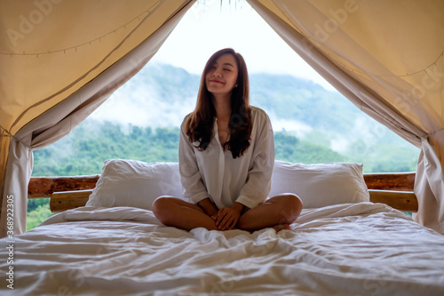 Portrait image of a young woman sitting on a white bed in the morning with a beautiful nature view outside the tent