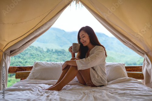 Portrait image of a beautiful young asian woman drinking coffee while sitting on a white bed in the morning with a beautiful nature view outside the tent