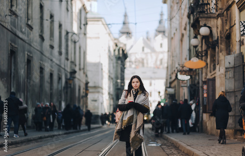A young girl in an autumn coat stands in the middle of the streets on a tram line and drinks coffee surrounded by passers-by. Lviv, Ukraine.