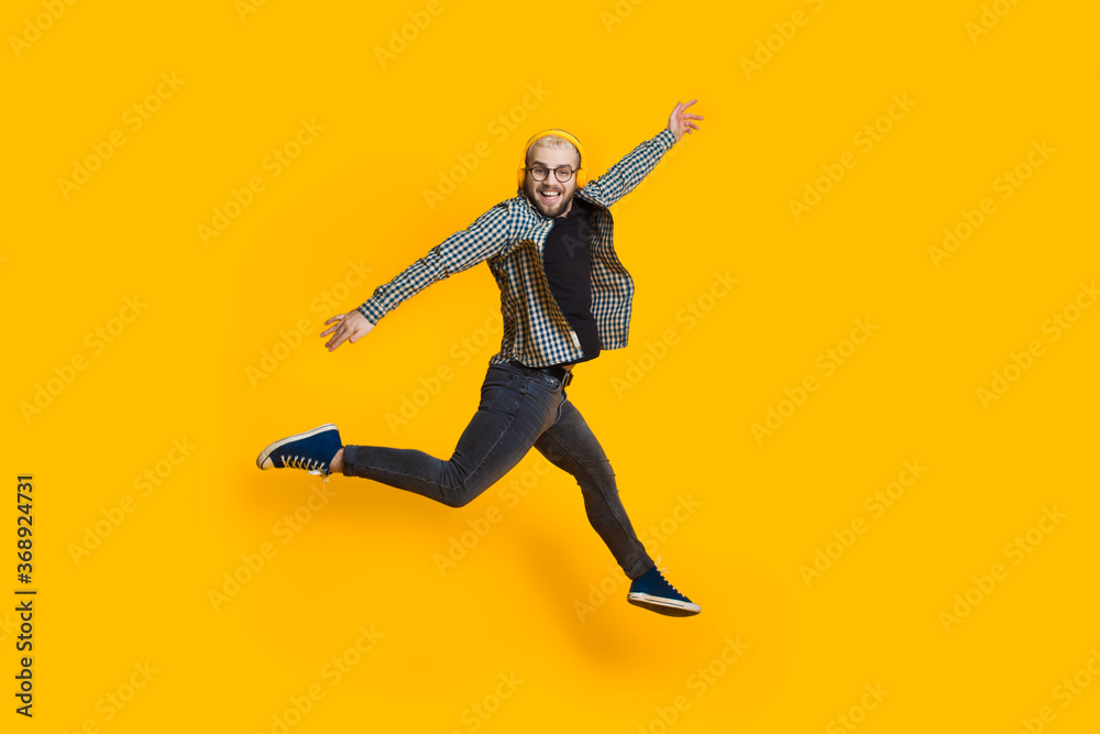 Jumping caucasian man with blonde hair and eyeglasses is listening to music on yellow studio wall