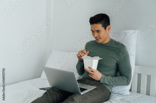 Portrait of Asian man eating takeout food watching laptop on bed. New lifestyle, food order from modern technology concept. photo