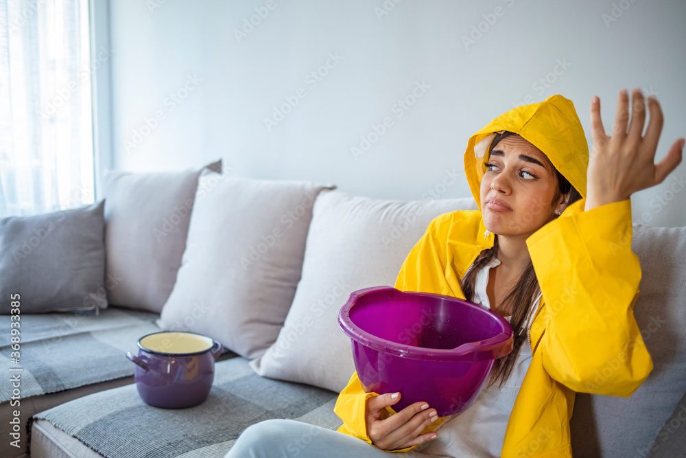 Woman collecting water leaking from the ceiling in home. Upset woman in raincoat calling maintenance service. Female having flood demage in house and using phone for help Woman collecting water leaking from the ceiling in home. Upset woman in raincoat calling maintenance service. Female having flood demage in house and using phone for help