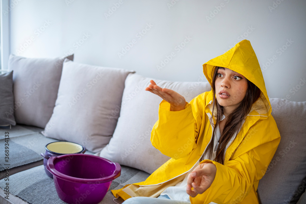 Frustrated homeowner watching ceiling leaks sitting on a couch at home. Time to call plumber. Shocked Woman in raincoat Looks at the Ceiling While Collecting Water Which Leaks Frustrated homeowner watching ceiling leaks sitting on a couch at home. Time to call plumber. Shocked Woman in raincoat Looks at the Ceiling While Collecting Water Which Leaks